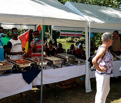 Auf diesem Foto vom Sommerfest der Kulturen in Fürstenau sieht man eine Szene von der kulinarischen Meile. In zwei weißen Pavillons sind Tische mit weißen Decken aufgestellt. Auf den Tischen stehen mehrere silberne Behälter mit Essen. Hinter den Behältern stehen drei junge Männer afrikanischer Abstammung. Der linke Mann trägt ein blaues T-Shirt und Handschuhe. Er trinkt gerade einen Schluck aus einer Flasche. Der junge Mann neben ihm trägt ein weißes ärmelloses Shirt mit einem grauen Balken auf der Brust, darüber ist mit dunkelblauer Schrift etwas geschrieben, was man nicht lesen kann. Der rechte Mann trägt eine schwarze Mütze mit rosa Schirm. Sein T-Shirt ist rot, schwarz und weiß gestreift. Er trägt ebenfalls blaue Handschuhe. Vor dem Pavillon rechts im Bildvordergrund stehen zwei Frauen, die sich anschauen und sich miteinander unterhalten. Die Frau, die etwas weiter links steht, trägt eine weiße Stoffhose und ein Blusentop mit Mustern. Sie hat graue kurze Haare und trägt eine Brille und steht seitlich zu der anderen Frau. Die andere Frau trägt eine blaue kurzärmlige Bluse und eine blaue Jeans. Vor ihrem Bauch trägt sie eine kleine schwarze Tasche.