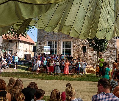 Beim Sommerfest der Kulturen in Fürstenau im Osnabrücker Land ist einiges los. Auf diesem Bild sieht man Kinder, die schätzungsweise zwischen sechs und sieben Jahre alt sind. Sie stehen aufgereiht wie ein Chor nebeneinander und schauen zum Publikum. Rechts in diesem Kinderchor steht ein kleiner Junge, der auf einem Klavier spielt. Hinter den Kindern sieht man einen Teil eines Steinwerks und zwei Fenster des Hauses mit weißen Sprossen. Vor dem linken Fenster steht ein weißes Banner, darauf ist mit weißer Schrift auf blauem Hintergrund „Freiwilligenagentur“ zu lesen. Darunter ist ein Bild mit Bäumen und einer grünen Blumenwiese zu erkennen, darauf steht mit weißer Schrift geschrieben: “Helfen macht glücklich”. Unter der Blumenwiese ist das Wappen von Fürstenau zu erkennen. Das Wappen ist rot und darauf ist eine graue Burg zu erkennen. Unter dem Wappen ist das Banner weiß und mit schwarzer Schrift steht geschrieben: Samtgemeinde Fürstenau. Rechts und links neben den Kindern steht jeweils ein schwarzer Lautsprecher. Links neben dem Steinwerk ist noch ein weiteres Haus zu erkennen. Dies ist aber deutlich kleiner und man sieht das Dach mit roten Dachpfannen. Vor dem kleineren Haus sieht man eine Familie stehen, ein kleiner Junge sitzt im Kinderwagen und dahinter stehen weitere Kinder. Das Foto ist aus dem Publikum gemacht worden. In der ersten Reihe sieht man Kinder und Eltern zur Bühne schauen. Im Bild sieht man sie nur von hinten. 